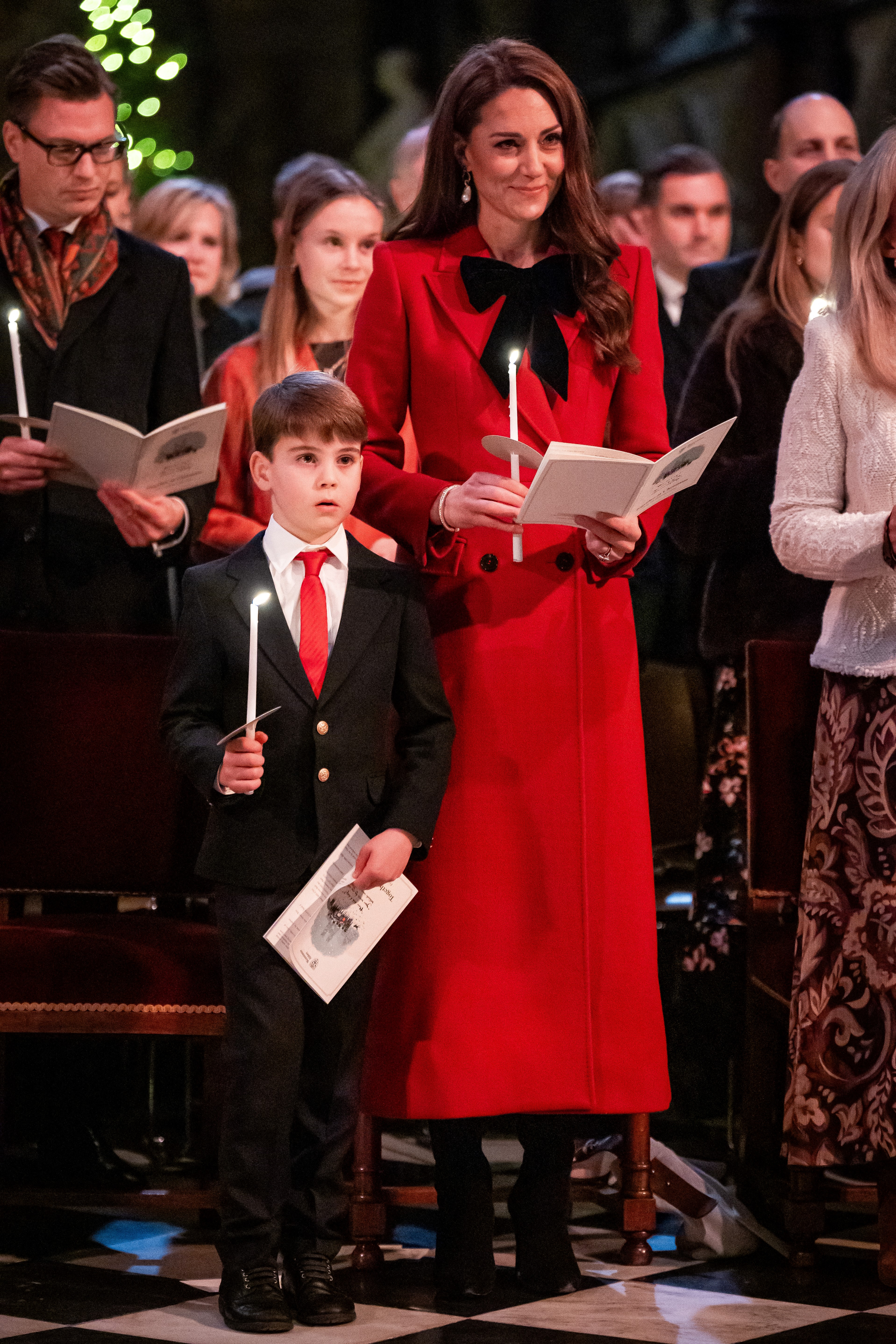 Kate Middleton holding a candle wearing a red coat next to Prince Louis