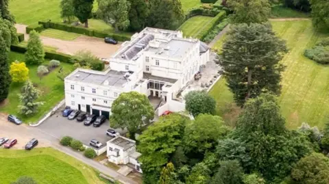 Shutterstock An aerial photograph of Royal Lodge, Prince Andrew's residence in Windsor, shows a large white building with green fields and trees surrounding it and a variety of cars parked outside it. File photo